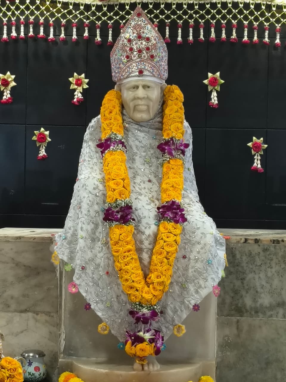 Close view of the murti's face and jeweled dome turban set with red gems, dressed in the white sequined shawl with a yellow rose and purple orchid garland