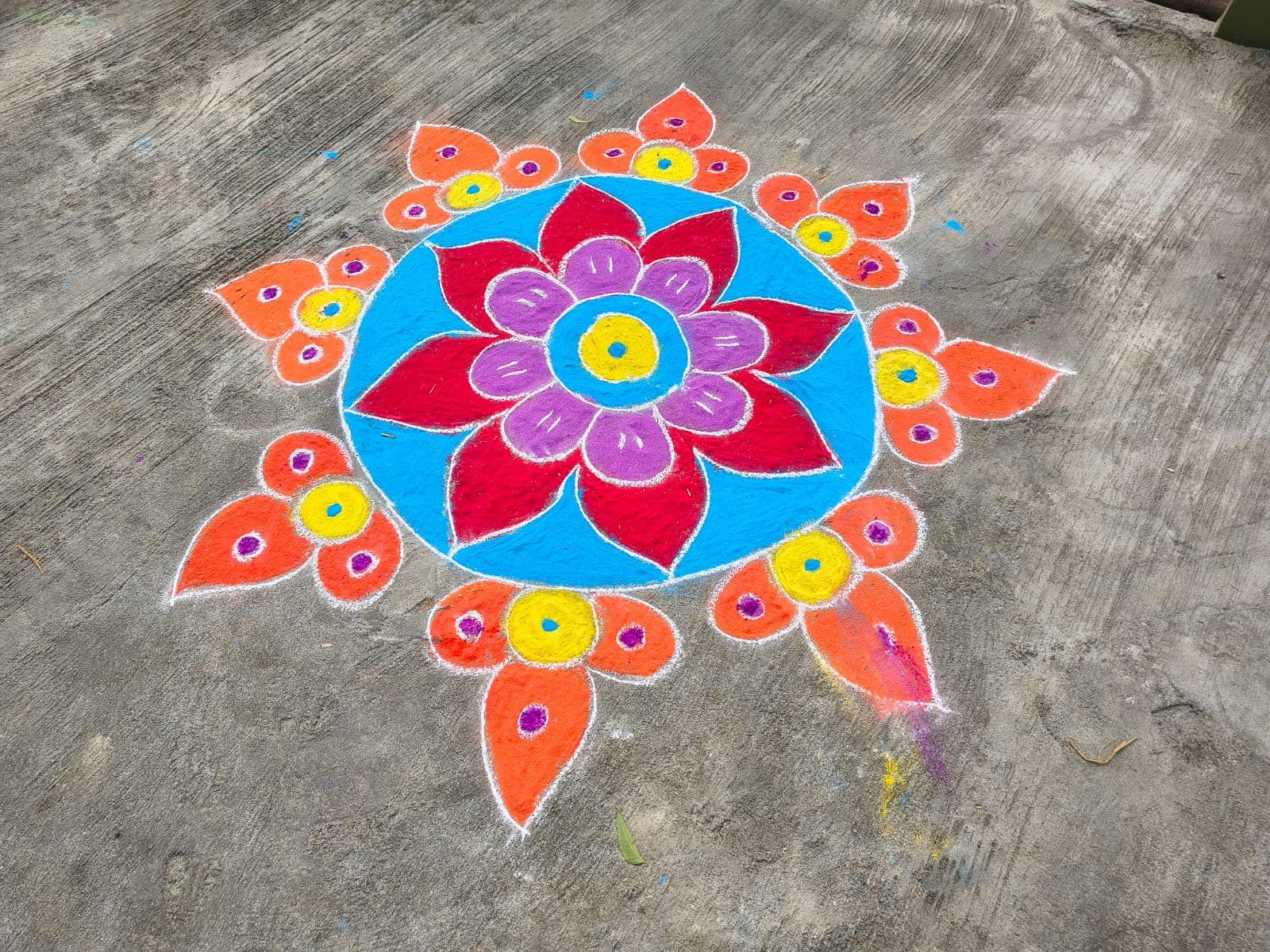 A rangoli of a red and purple lotus flower on a blue circular field, bordered by orange petal points