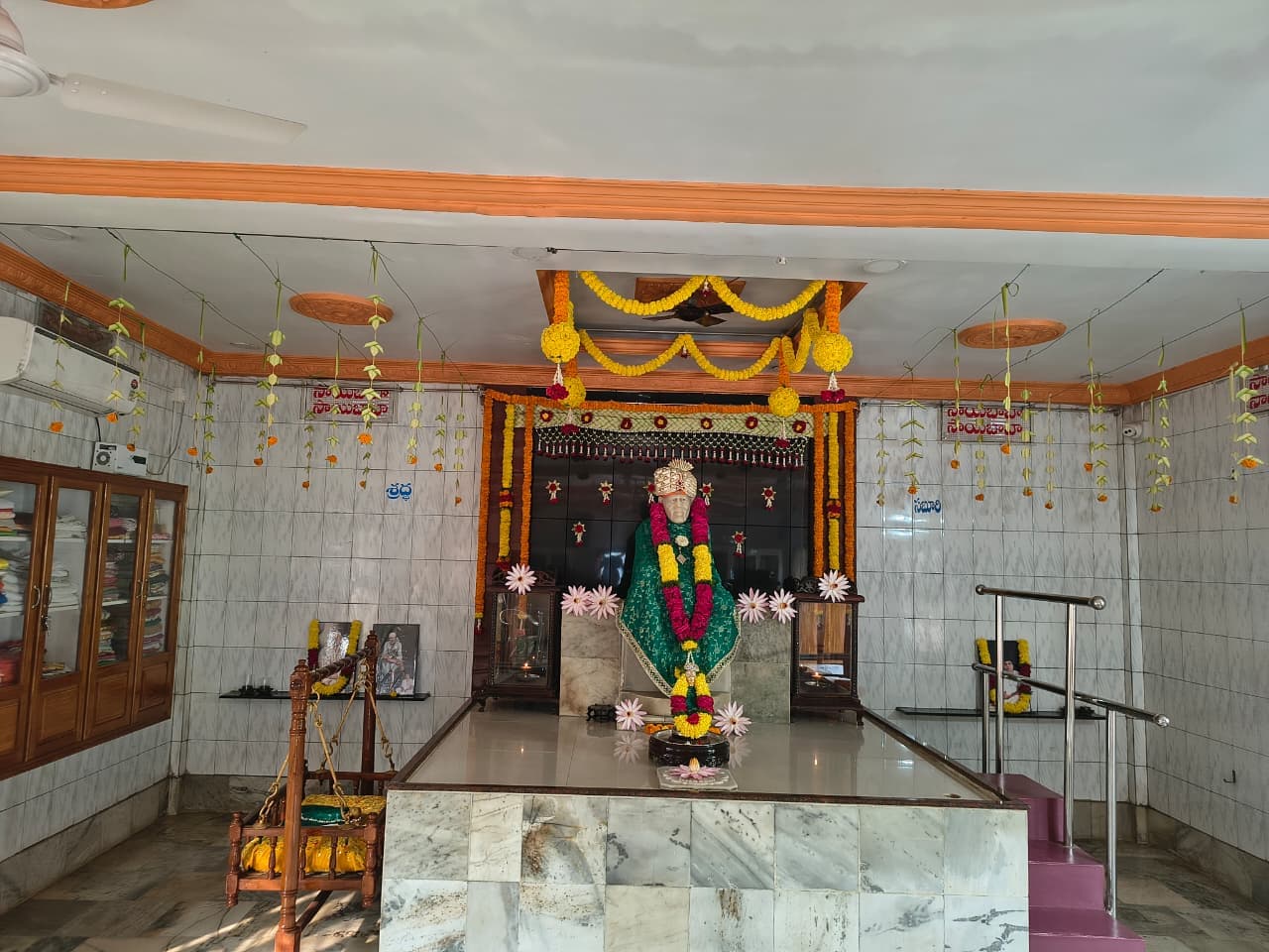 Wide view of the sanctum, the murti bust dressed in a green shawl and jeweled turban with an "ॐ" mark, beneath marigold garlands, with "శ్రధ్ధ" and "సబూరి" lettered on the side walls