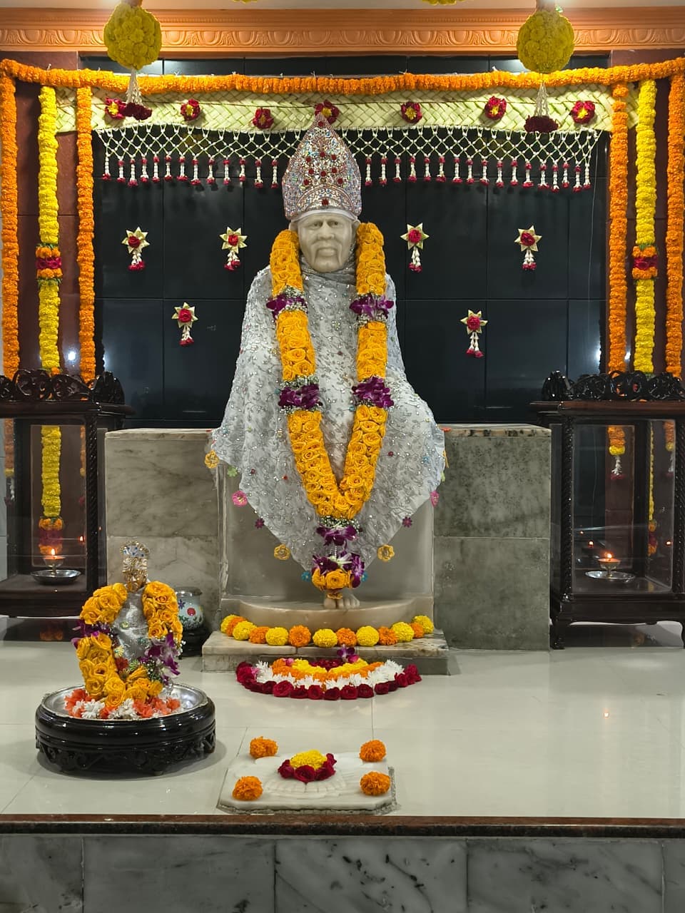 Full view of the murti in the white sequined shawl and jeweled turban, the small utsav murti and marigold and rose offerings arranged at the base