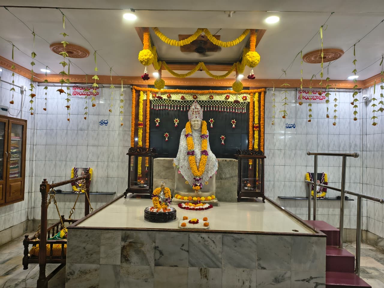 Wide view of the sanctum, the murti now re-dressed in a white sequined shawl with an orange, yellow, and purple flower garland, a small utsav murti and floral offerings at the base