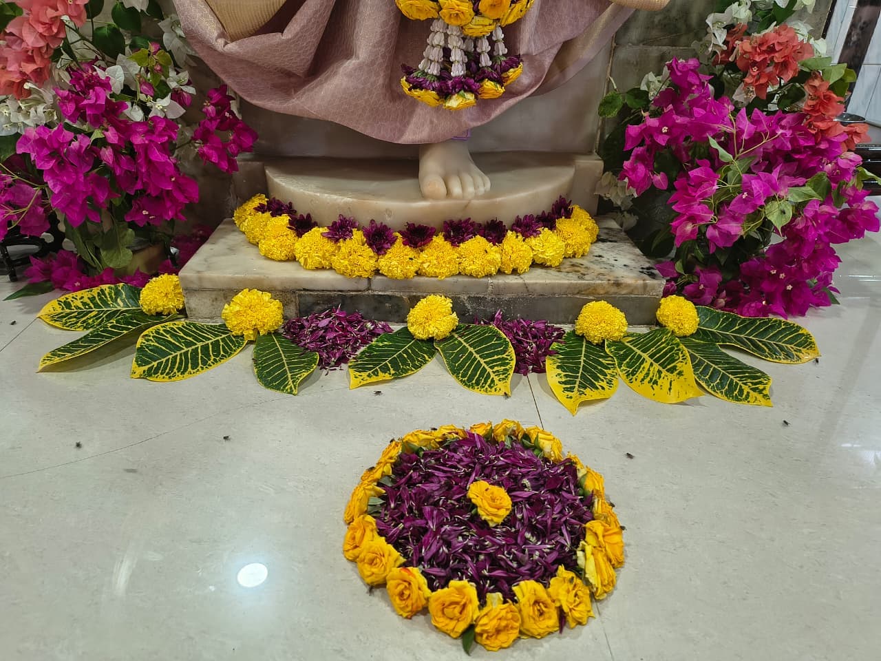A floor arrangement of marigold flowers, purple petals, and decorative leaves laid at the murti's feet