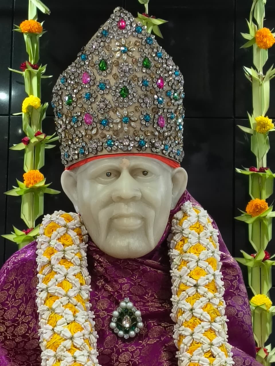 Close view of the murti's face and the jeweled crown set with colored stones, worn with the purple shawl