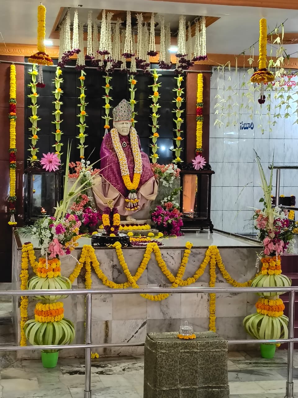 The murti dressed in a purple and gold shawl and a jeweled crown, framed by hanging white and purple garlands and pink and white flowers at the altar