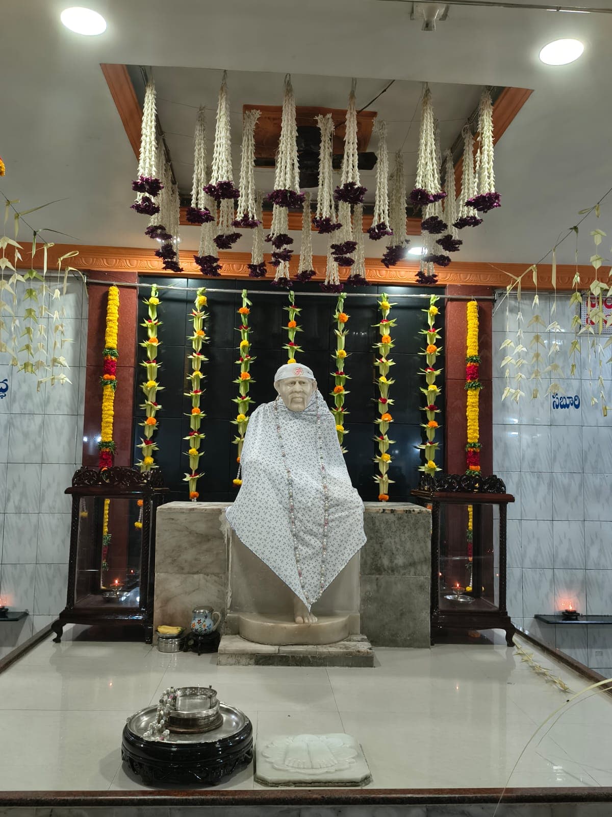 The murti dressed in a white floral-print shawl marked with an "ॐ" on the head-cloth, wearing a long beaded mala, framed by hanging white and purple flower garlands