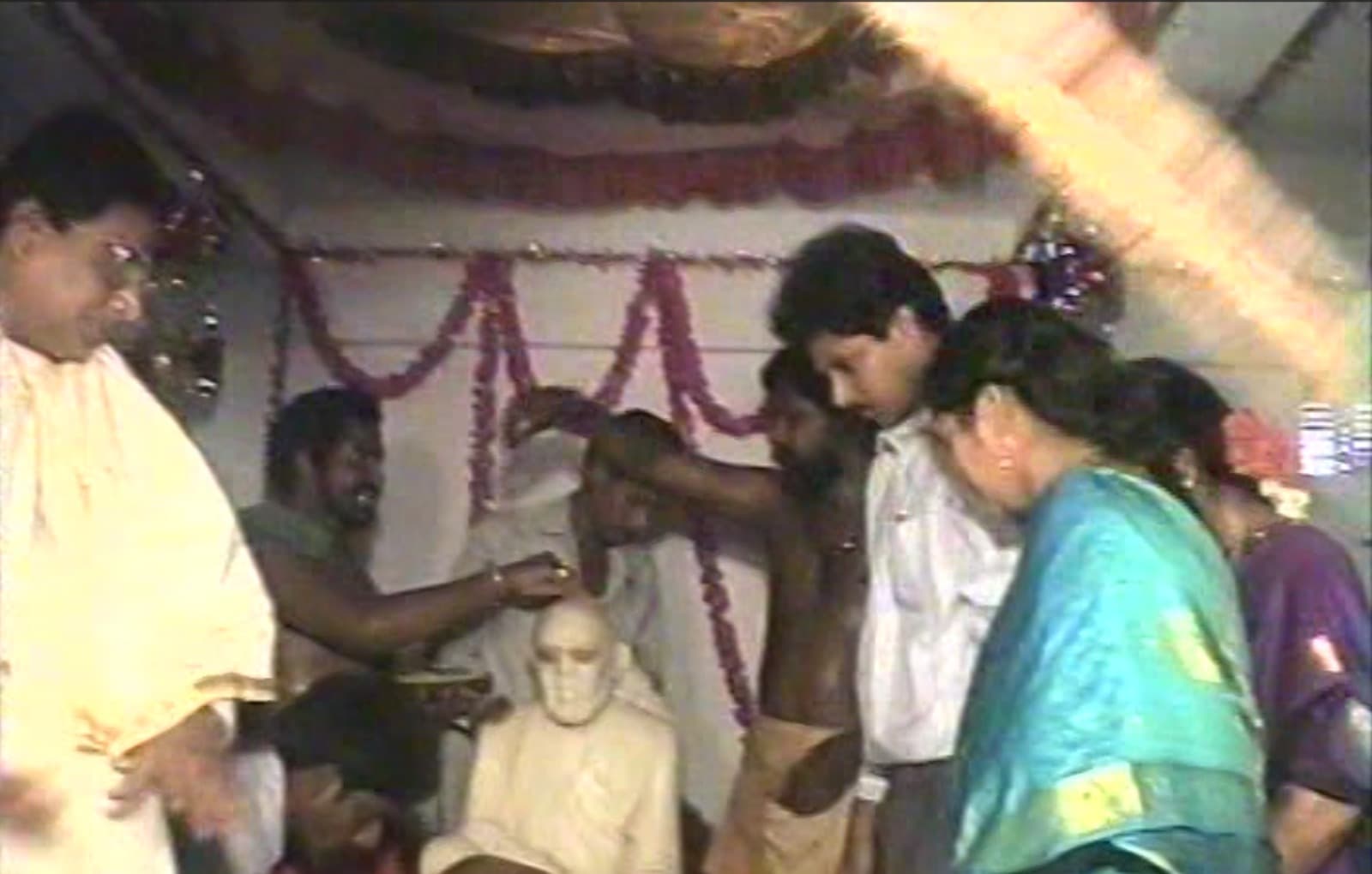 Archival 1997 photo of priests performing the abhishekam on the newly consecrated murti, anointing its head while devotees gather closely around under a garland-decorated ceiling