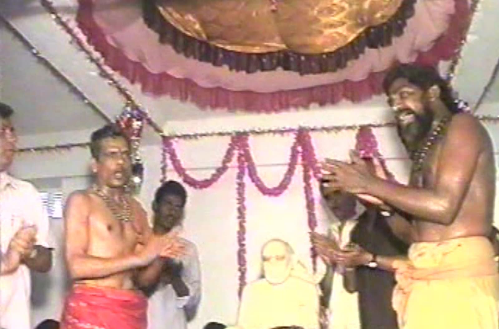 Archival 1997 photo of two men exchanging a namaste greeting beside the newly consecrated murti, framed by garlands hanging from the ceiling, devotees looking on