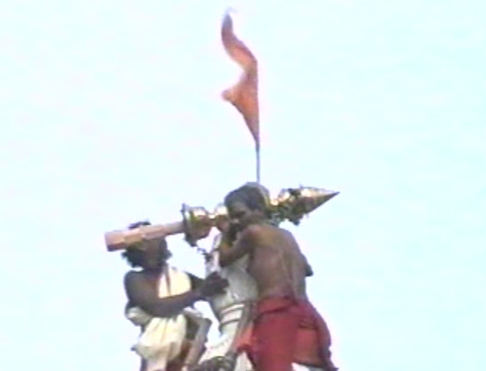 Archival 1997 photo of two men hoisting a flag atop the temple's gold kalash finial against a bright sky, part of the consecration ceremony