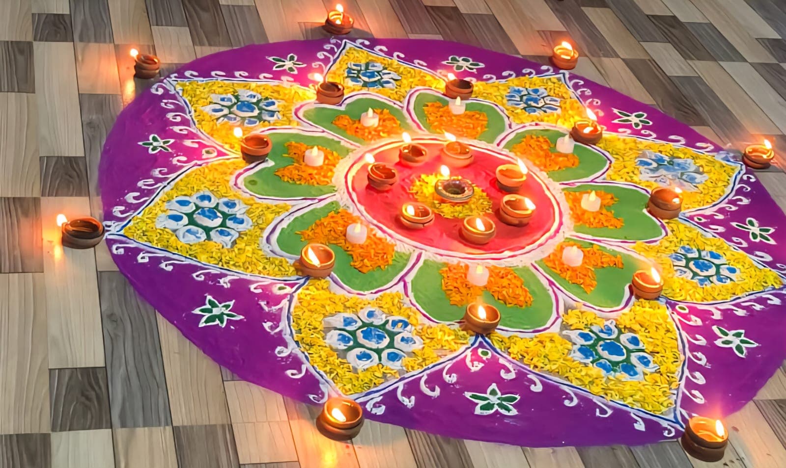 A large rangoli floor design in purple, yellow, and green, surrounded by lit oil lamps for a temple celebration