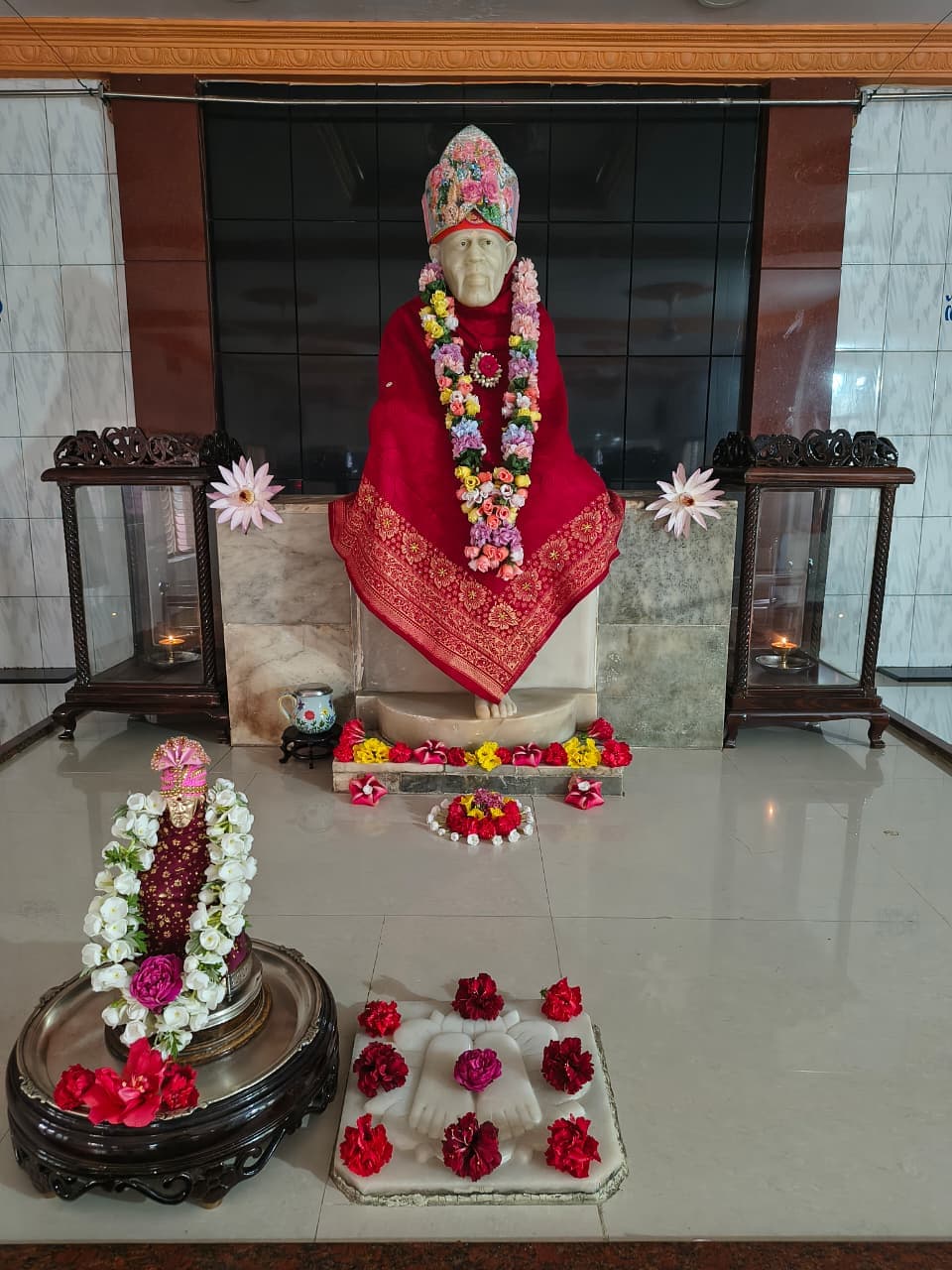 Murti in a red silk robe with a floral crown, a multicolor garland, and a small companion murti in front