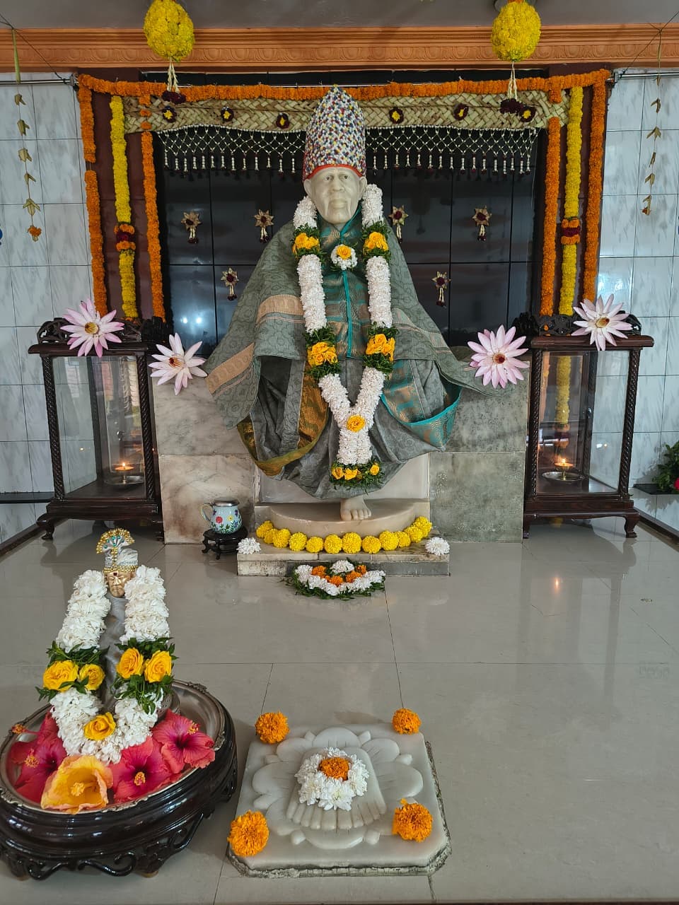 Murti in a sage-green and gold brocade robe under a marigold garland arch