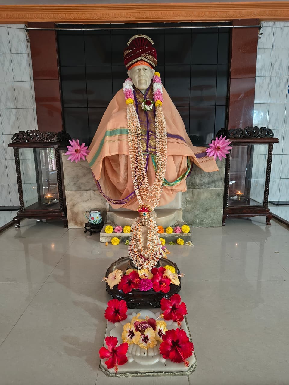 Murti in a peach-and-gold brocade robe with a maroon velvet turban, a small companion murti with hibiscus offerings in front