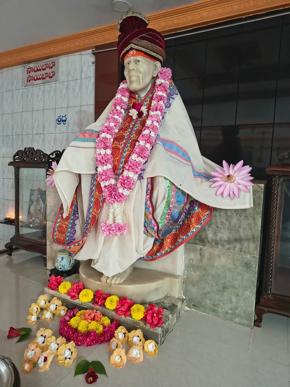 Murti in a cream robe with a maroon turban and a pink-white-red garland, against a fairy-light and floral backdrop