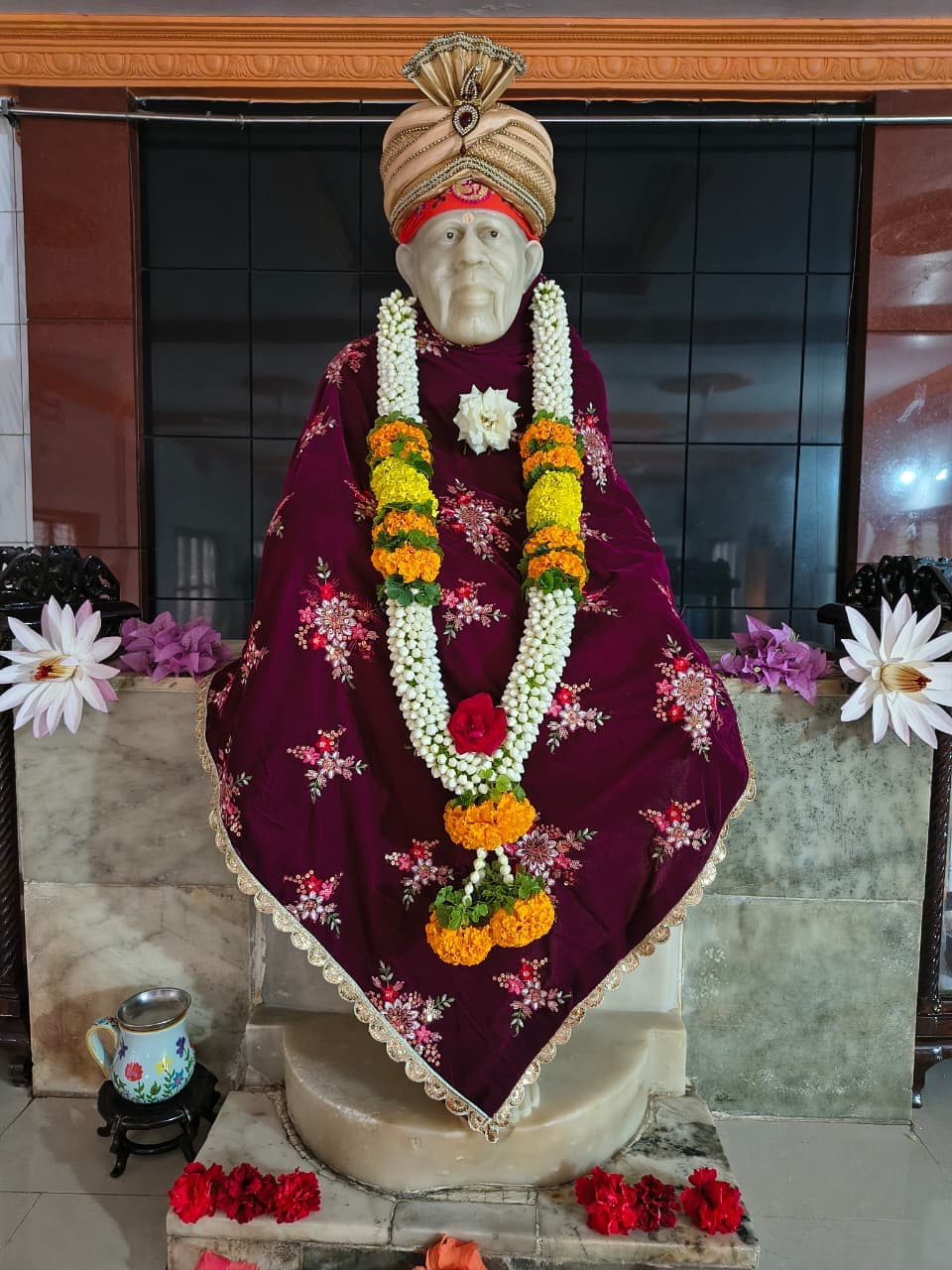 Murti in a royal blue robe with a silver floral pattern, a silver-and-purple garland, and a small companion murti in front