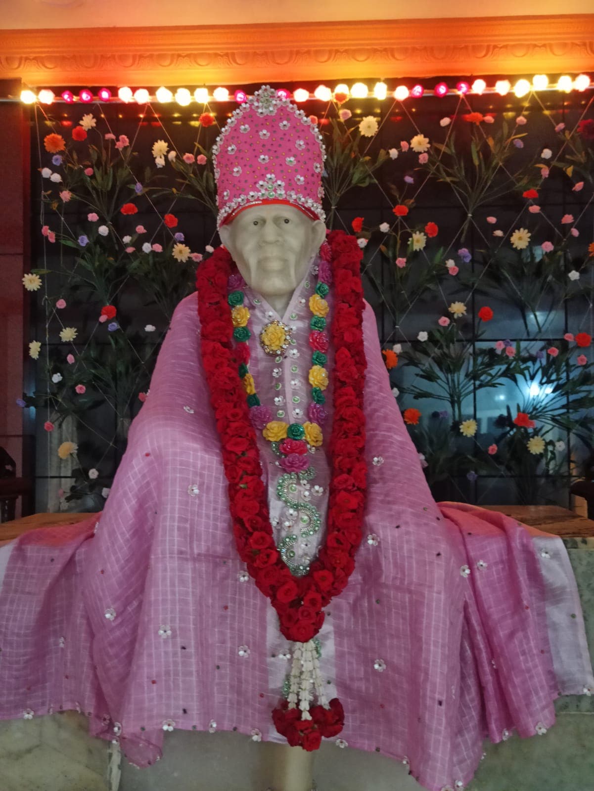 Murti in a pink checked silk robe with a jeweled crown and a red rose garland, against a fairy-light backdrop