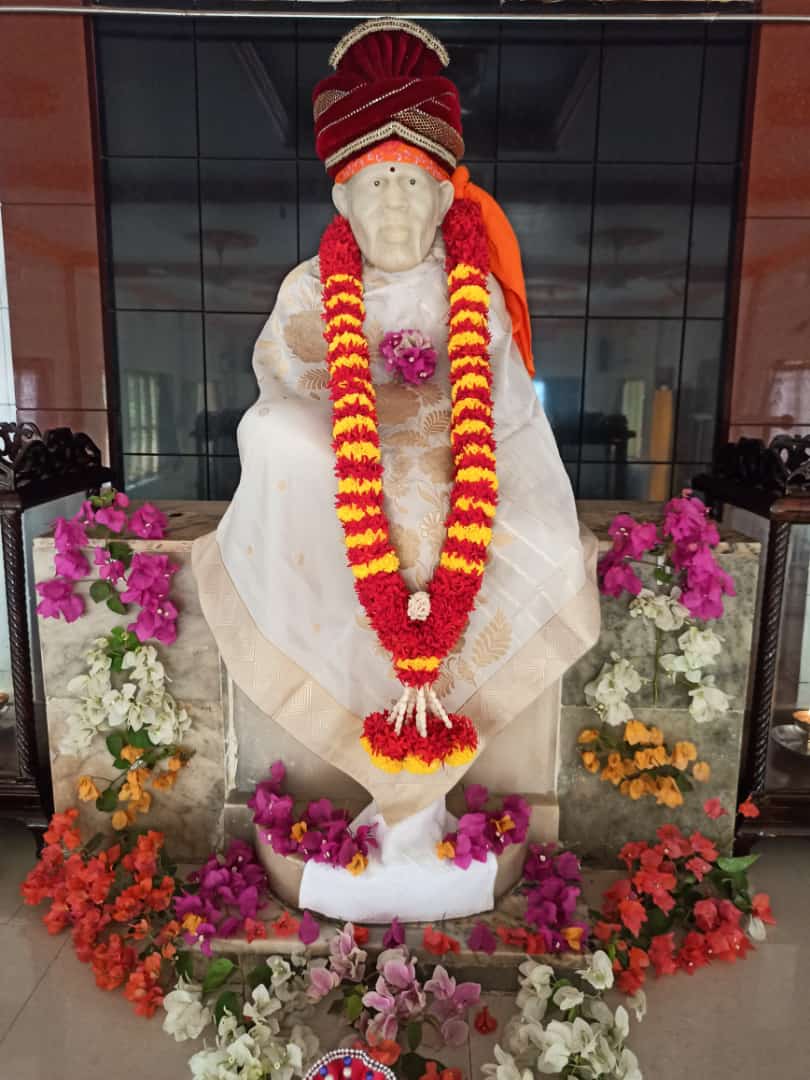 Murti in a white-and-cream robe with a maroon turban and a red-and-yellow striped garland