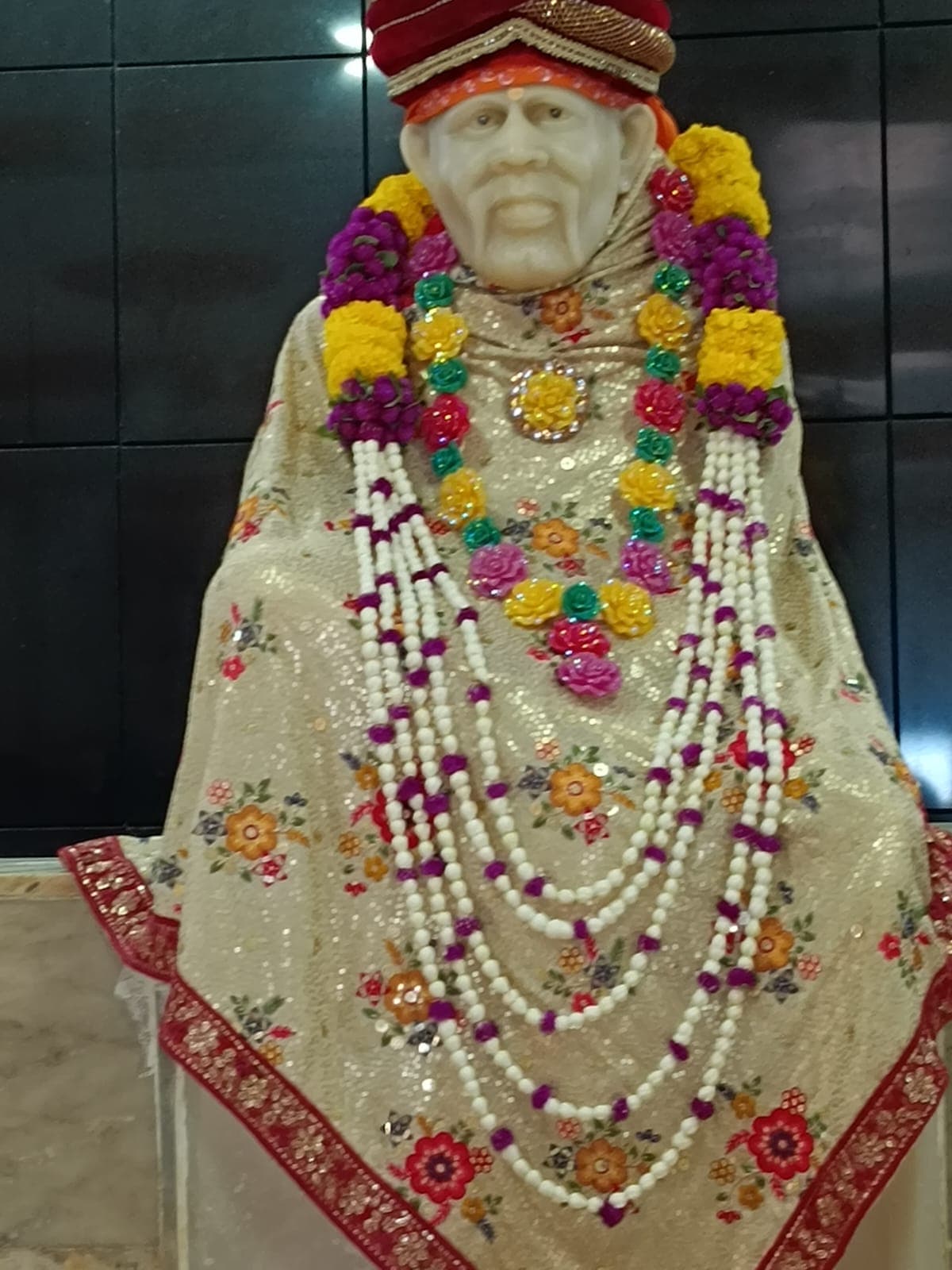 Murti in a gold-and-cream floral brocade robe with a multicolor rose garland over long beaded necklaces
