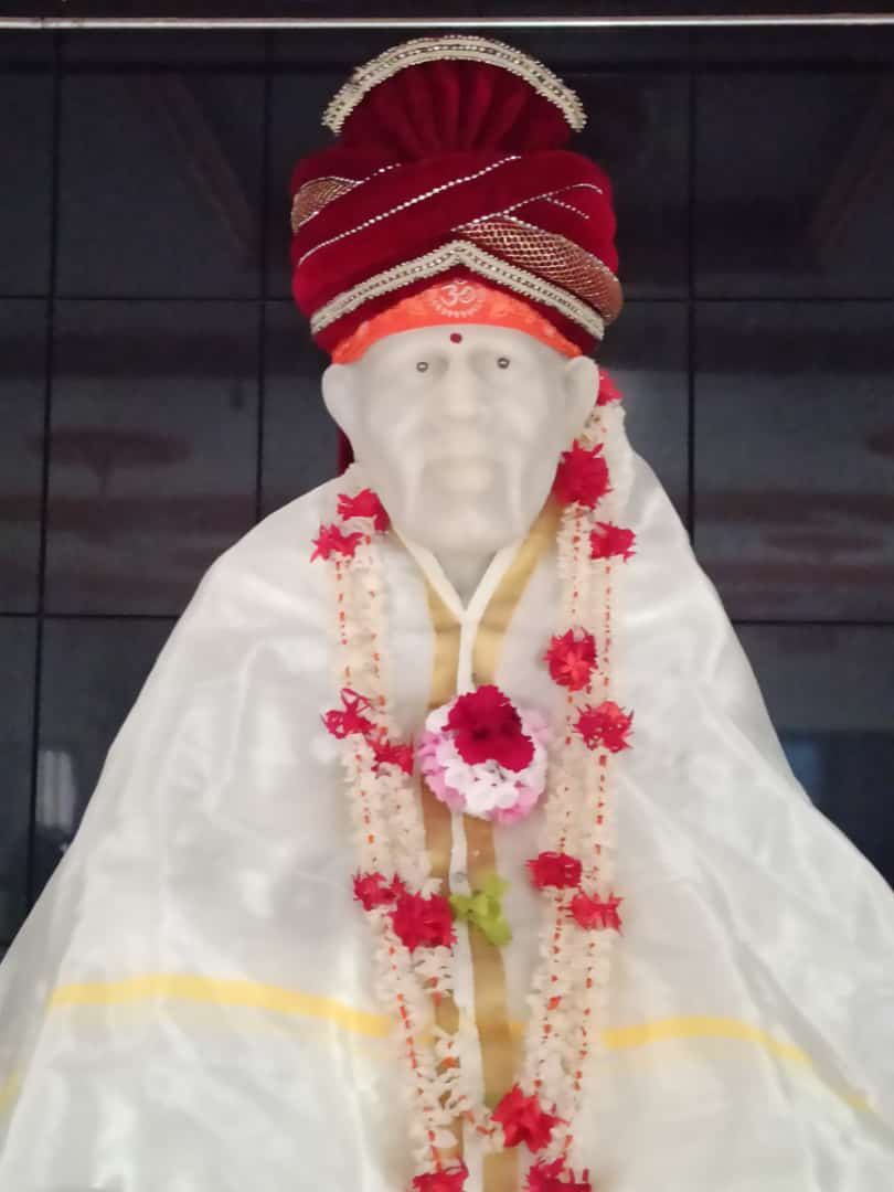 Murti in a sheer white robe with gold trim, a maroon turban, and a red-white-pink floral garland