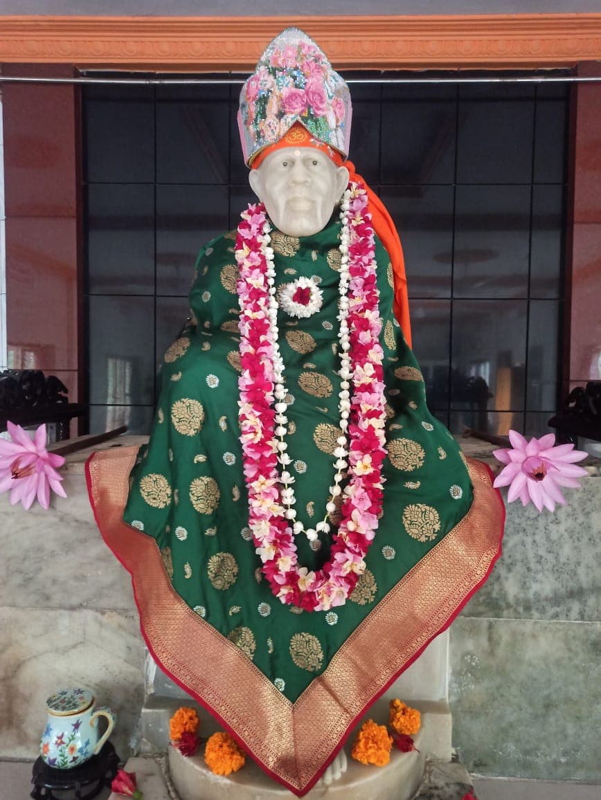Murti in a dark green and gold brocade robe with a beaded floral crown and a pink-white-red garland