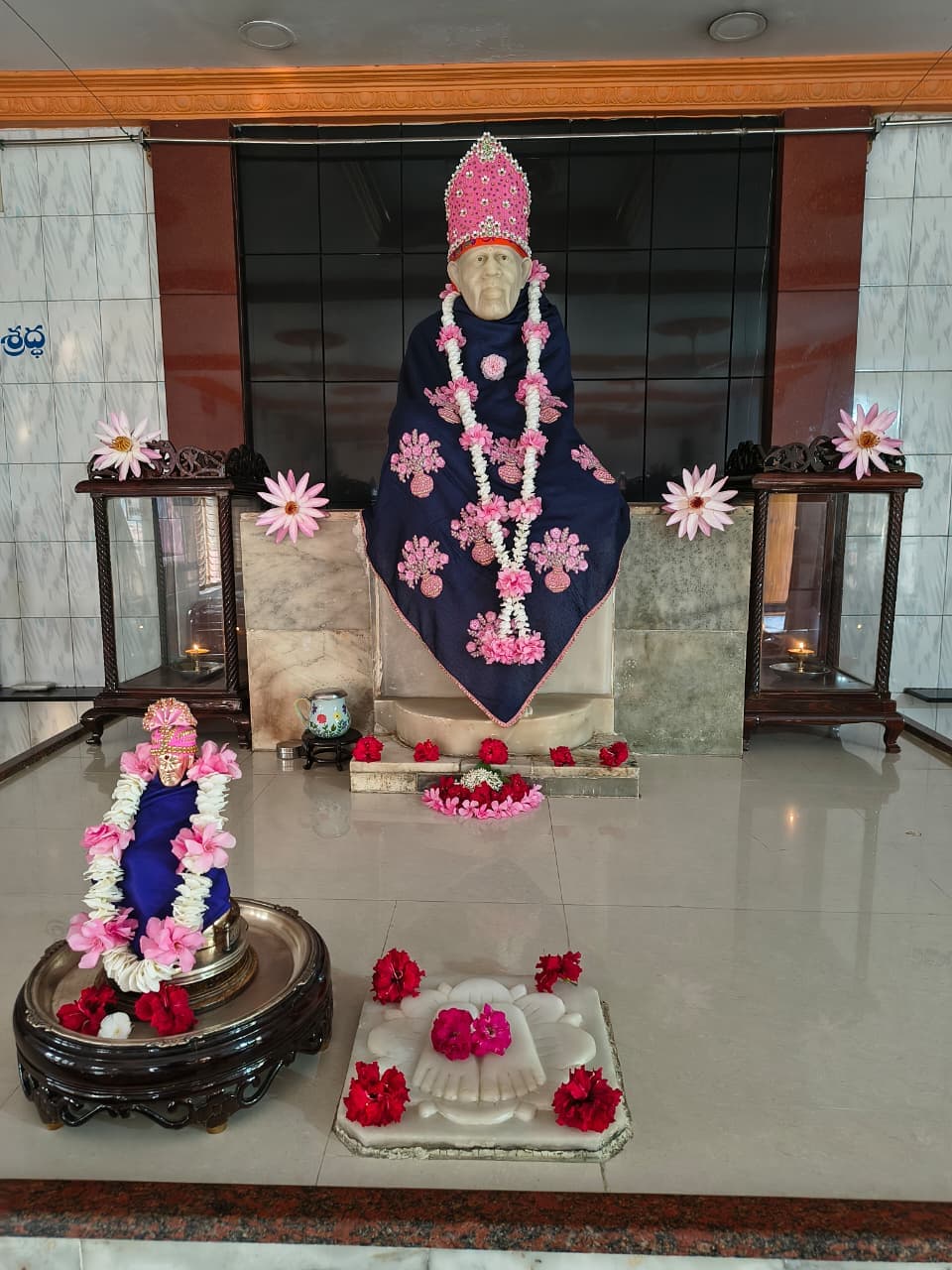 Murti in a navy blue robe with pink floral medallions, a jeweled pink crown, and a small companion murti in front