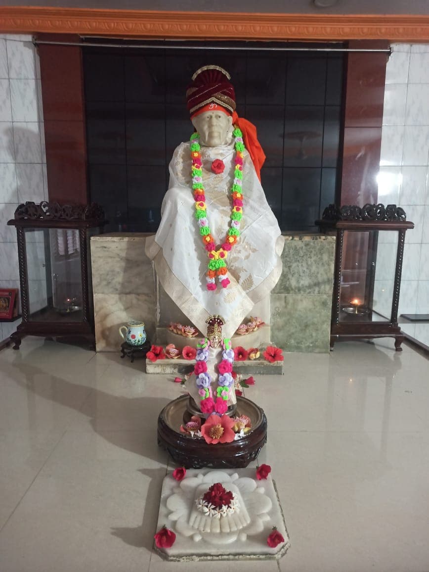 Murti in a cream-and-white robe with a maroon turban and a multicolor garland, a small companion murti with pink flowers in front