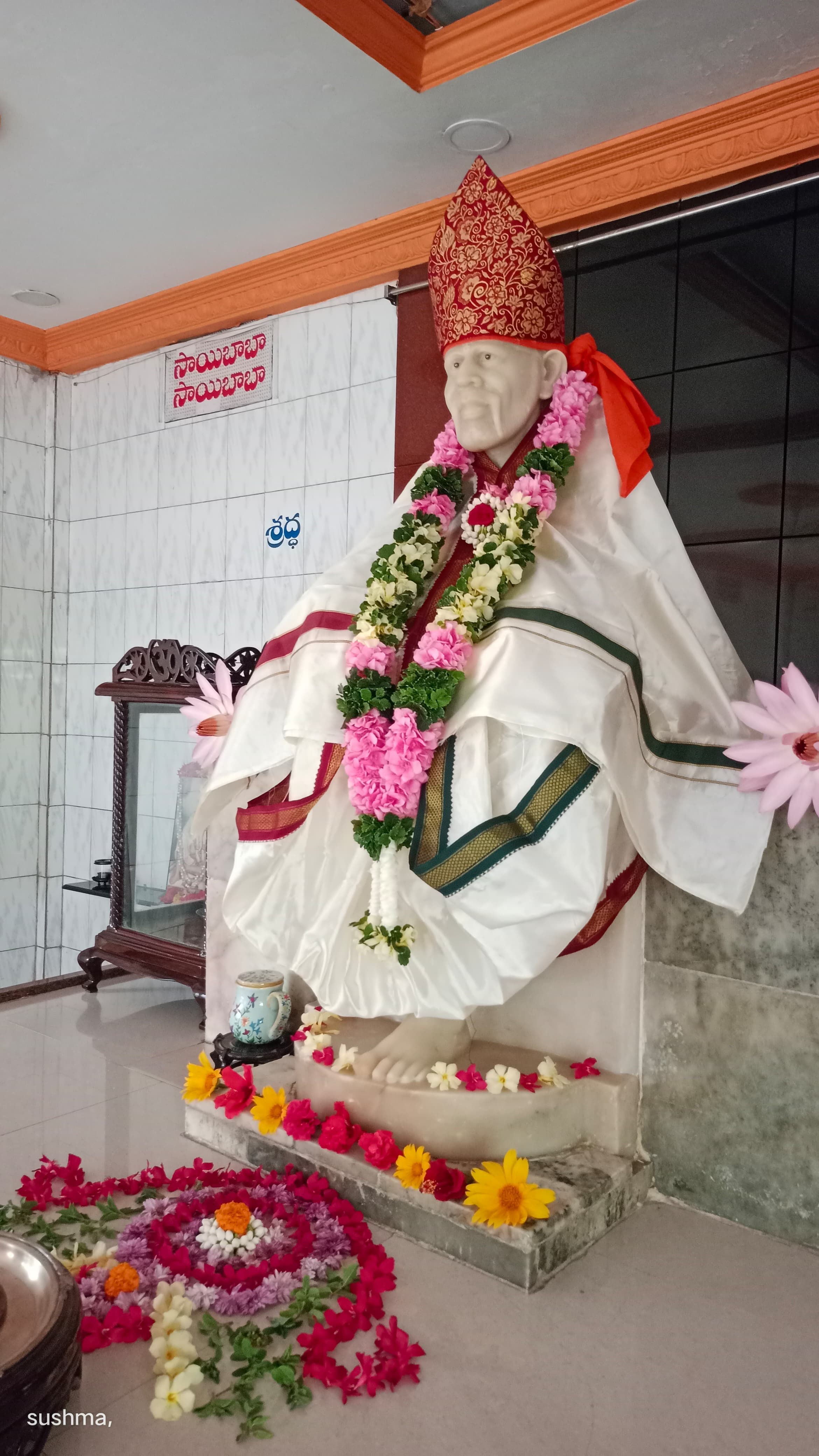 Murti in a white-and-green silk robe with a red floral-patterned crown and a pink-white-green garland