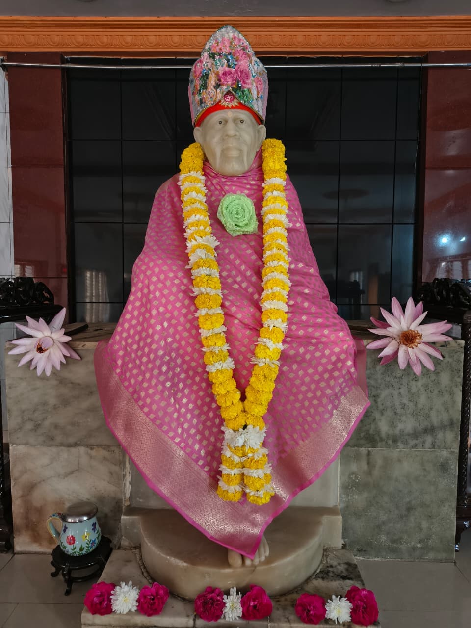 Murti in a pink diamond-pattern silk robe with a floral jeweled crown, a marigold garland, and a green fabric-rose brooch