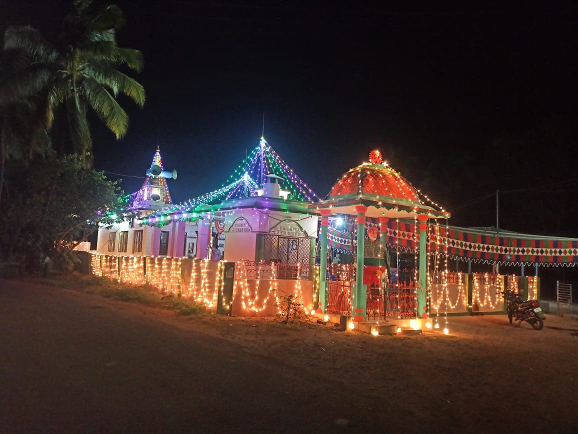 Temple exterior at night, decorated with string lights and a small lit mandapam, palm tree in the foreground
