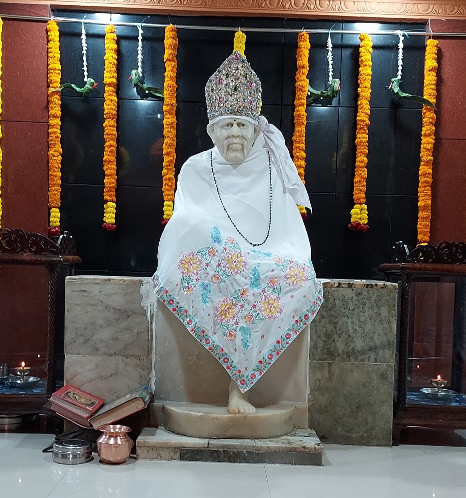 Murti in a white floral-embroidered shawl and rudraksha necklace, dark tiled backdrop with marigold garlands