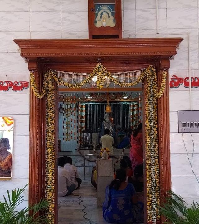 View into the sanctum through the carved wooden entrance doorway, garlanded with marigolds, with "Sai" and "Baba" lettered on the wall on either side, devotees seated inside