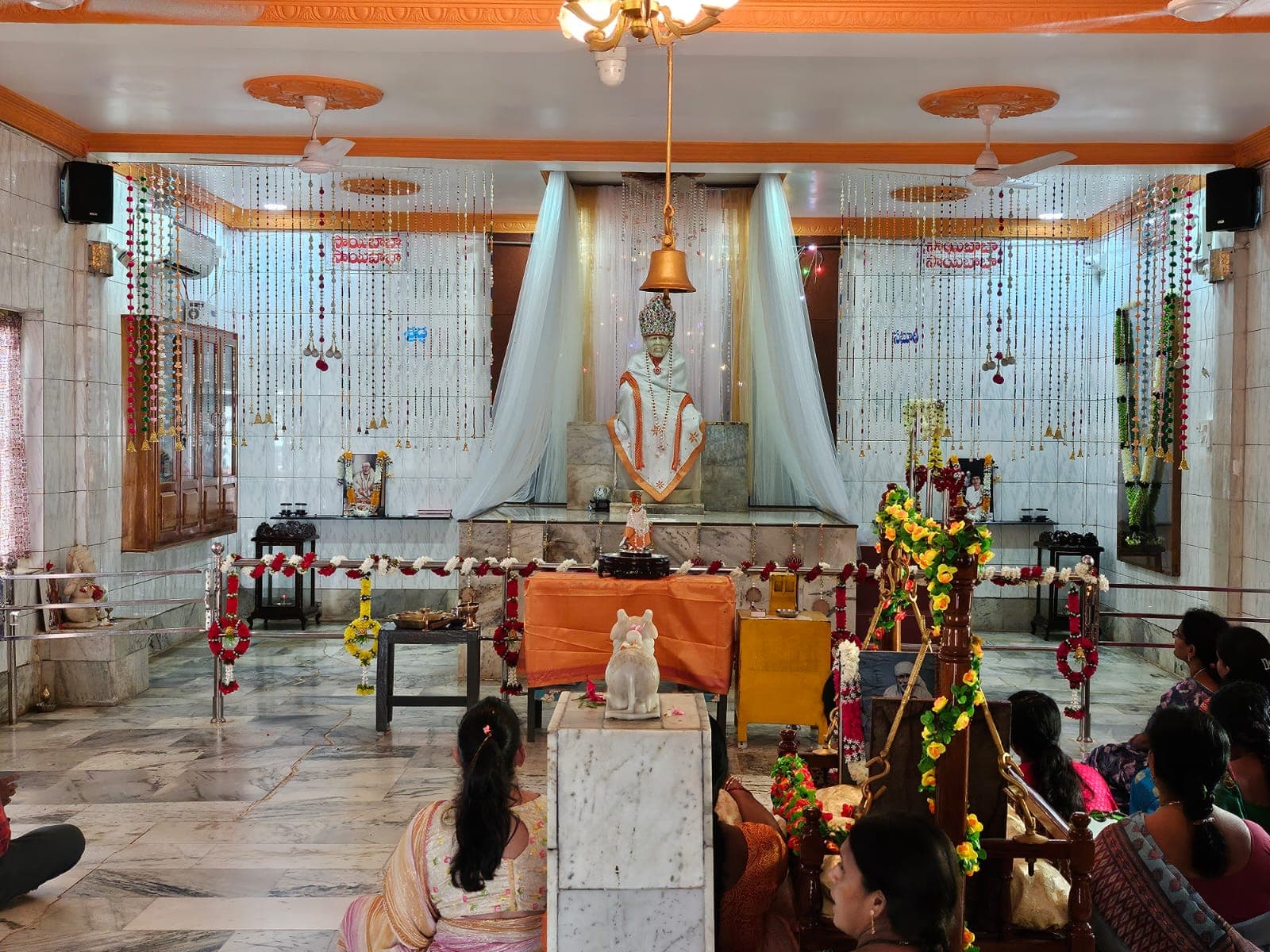 Sanctum interior view of the main Sai Baba murti, draped in white and orange cloth with a jeweled crown and marigold garlands, framed by sheer curtains and bead-and-bell toranams, with devotees seated before the altar