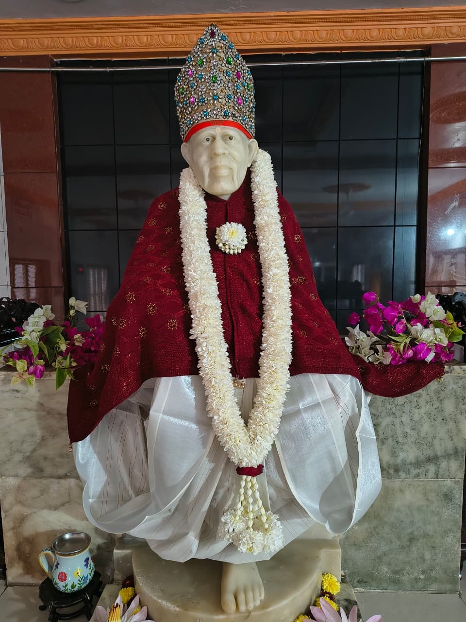 The murti in a maroon shawl and jeweled crown, garlanded in white