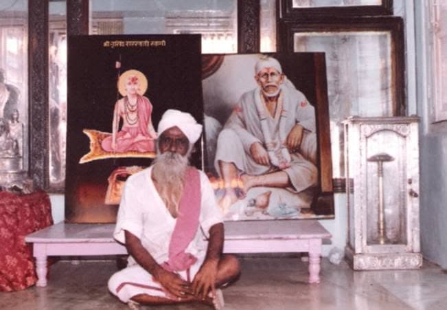 Sri Sivanesan Swami seated before framed paintings, including one of Sai Baba