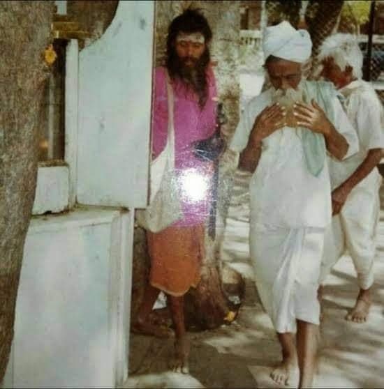 An older photo of Sri Sivanesan Swami standing beside a white shrine wall, an elderly devotee with hands folded in prayer walking past